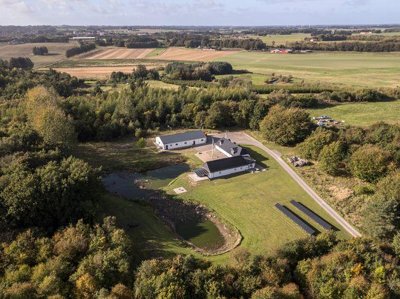 Luftfoto af hvide bygninger med mørke tage, en lille sø, plæne og solpaneler på jorden, omgivet af skov og åbne marker.
