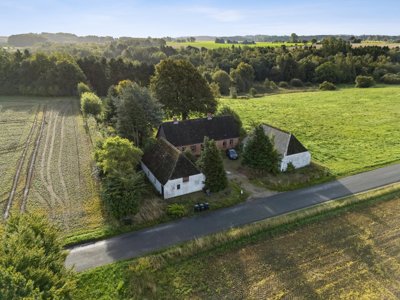 Aerial view of a white and red farmhouse with dark roofs and an outbuilding by a road, surrounded by fields, trees and forest.