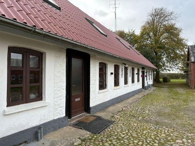 Wit gepleisterd gebouw met rood pannendak, dakramen en bruine deuren en ramen aan een keistenen erf met grote bomen.