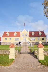 Geel hoofdgebouw met rode pannendaken en dakkapellen, grindoprit met hek en gele pilaren, gazons en een vlaggenmast.