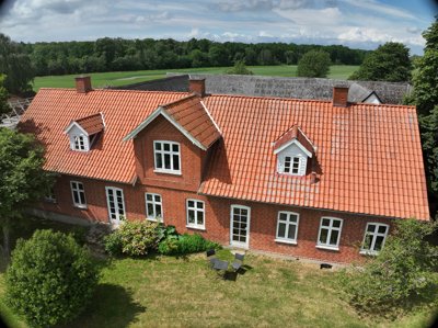 Set oppefra: rødt murstenshus med røde teglsten, kviste og have med plæne og buske; marker og skov i baggrunden.
