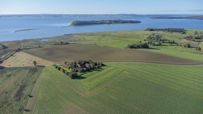 Luftfoto af en lille gård mellem træer omgivet af marker, med en fjord og en skovklædt ø i baggrunden.